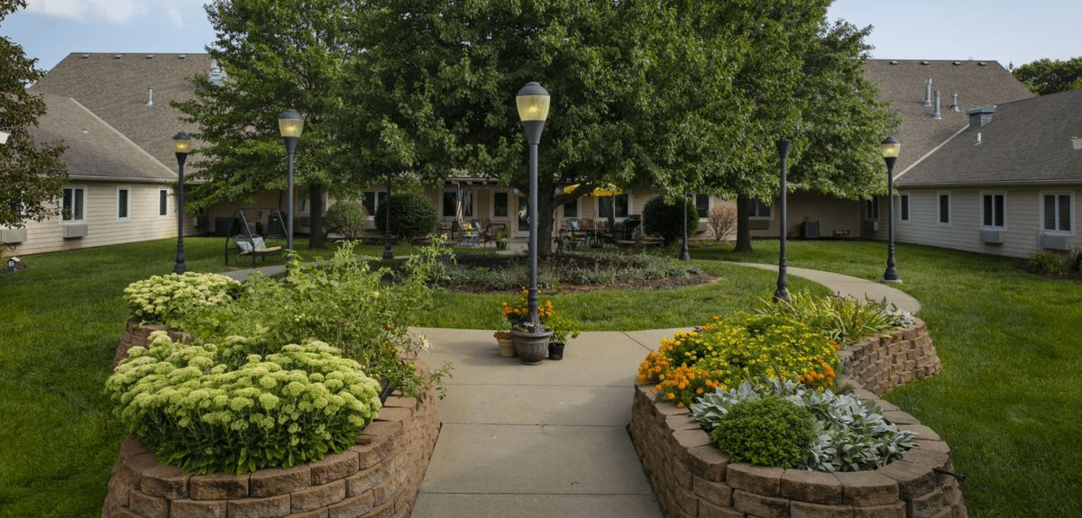 Pathway through lush lawn at Homestead of Shawnee senior living community with bench and potted plants.
