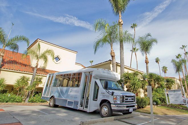 Senior resident boarding a minibus at Villa De Anza Assisted Living facility, surrounded by lush gardens.