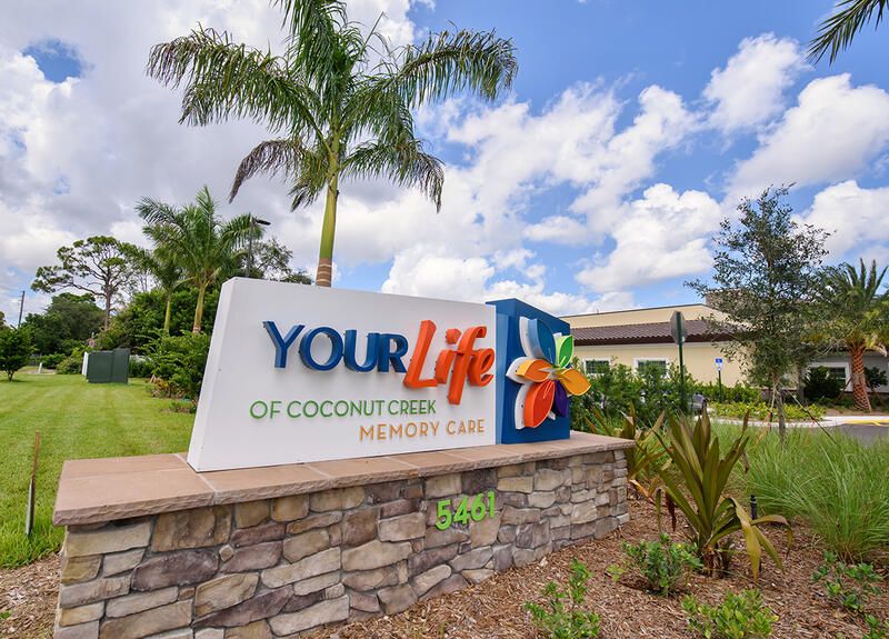 Summer view of The Monarch senior living community in Coconut Creek, featuring lush vegetation.