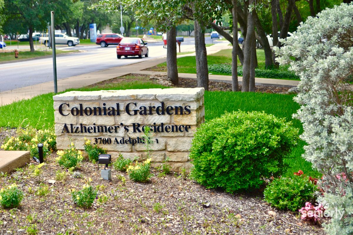 Seniors enjoying a peaceful walk in the lush gardens of Colonial Gardens of Austin with pathways, trees, and vehicles nearby.