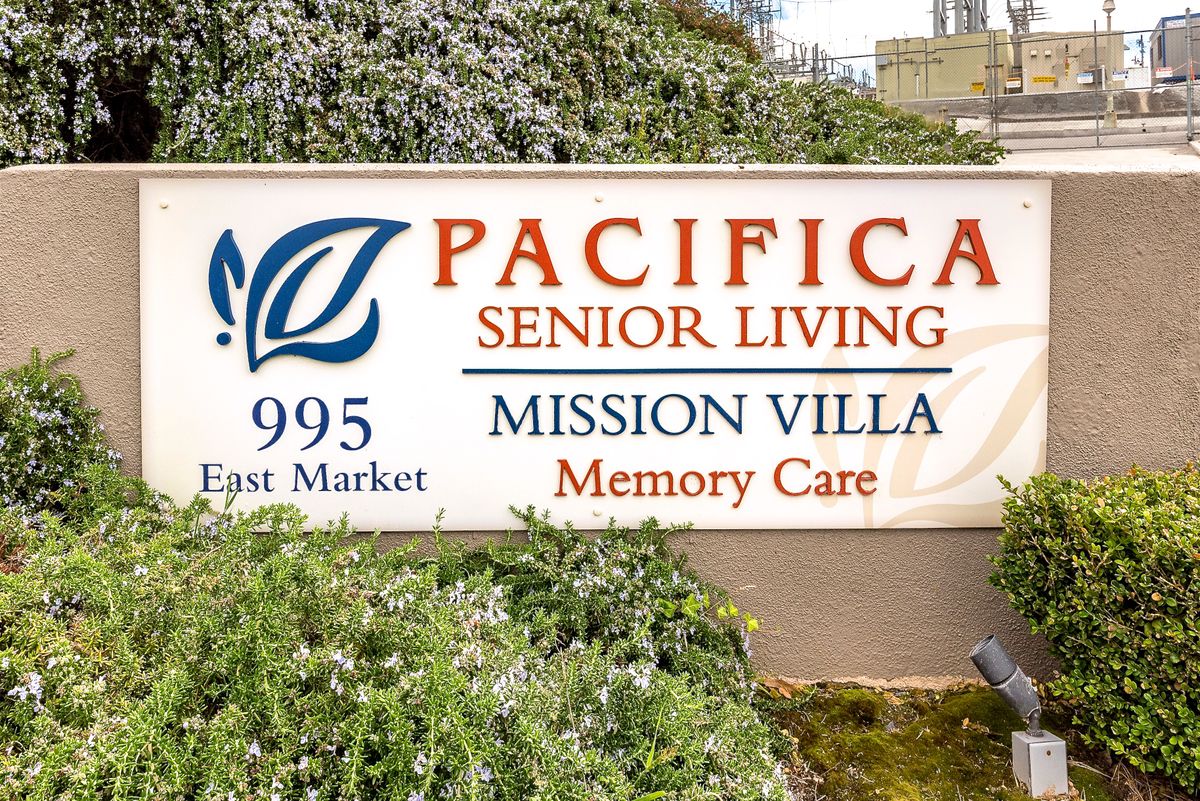 Sign for Pacifica Senior Living Mission Villa amidst lush vegetation outdoors.