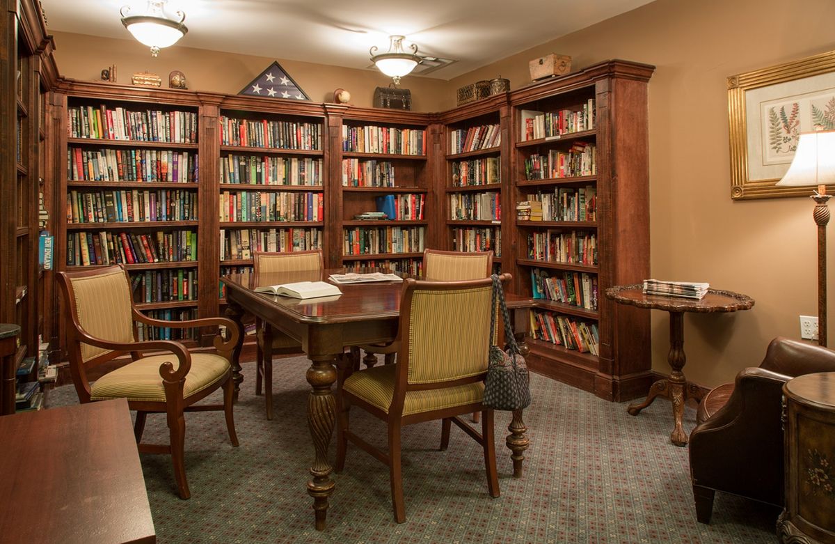 Senior living community library in Dedham with chairs, bookcases, table, lamp and accessories.