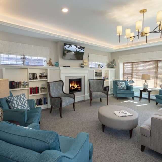 Senior living room at Compass On The Bay with cozy furniture, decor, fireplace, and electronics.