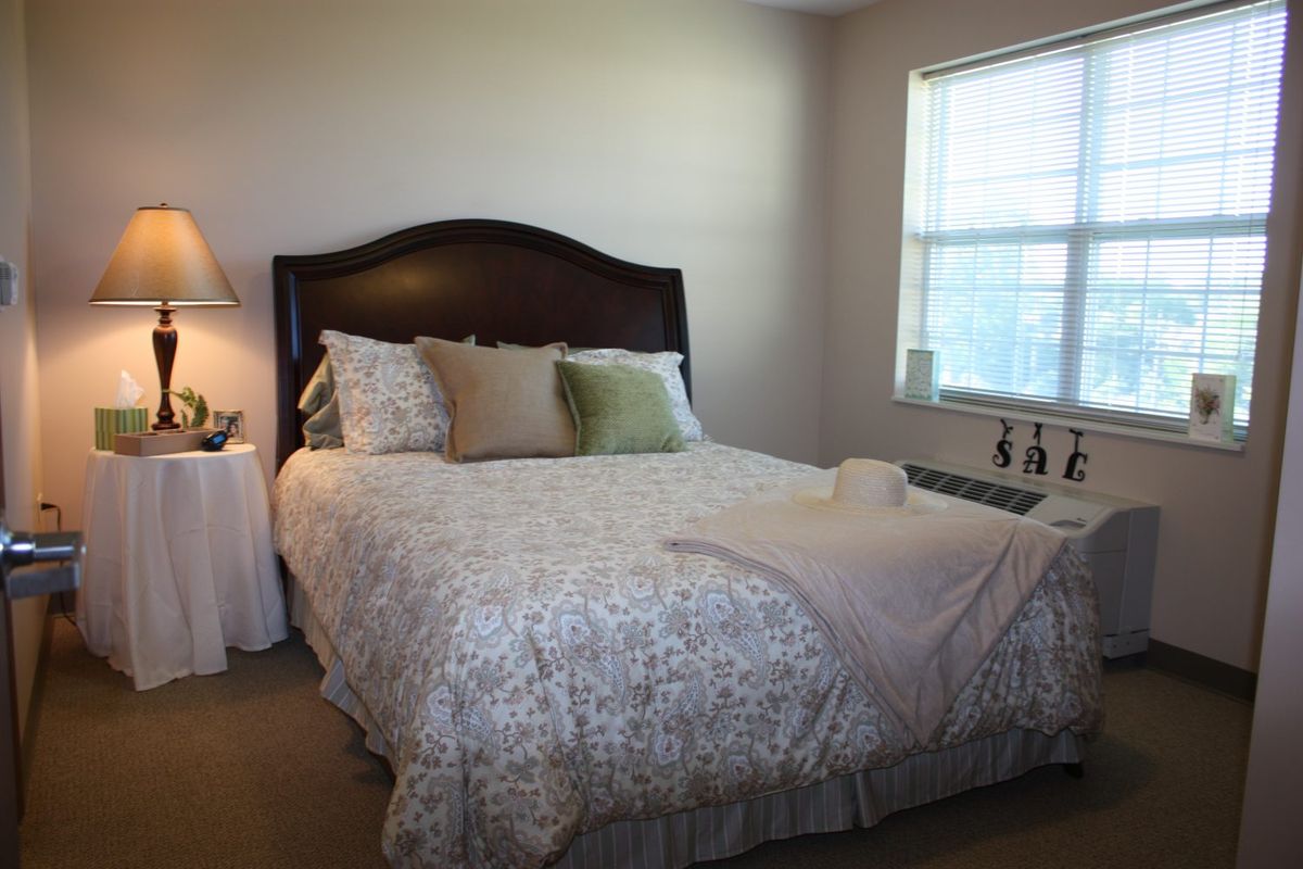 Interior view of a bedroom at Montclare Senior Living featuring a bed, table lamp, and home decor.