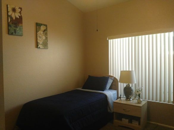 Corner bedroom in Rhodas Assisted Living Home featuring a bed, table lamp, indoor plant, and home decor.