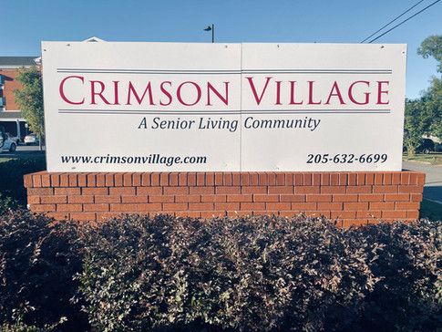 Advertisement sign for Crimson Village senior living community, surrounded by lush vegetation.