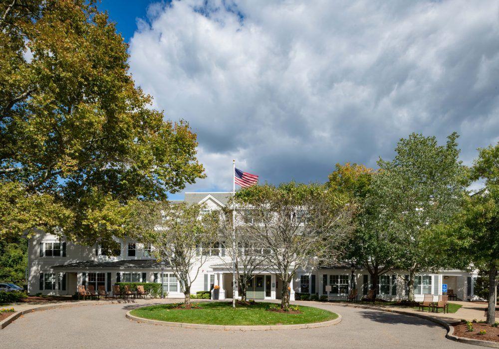 Exterior view of The Linden at Dedham senior living community with lush vegetation and cars.
