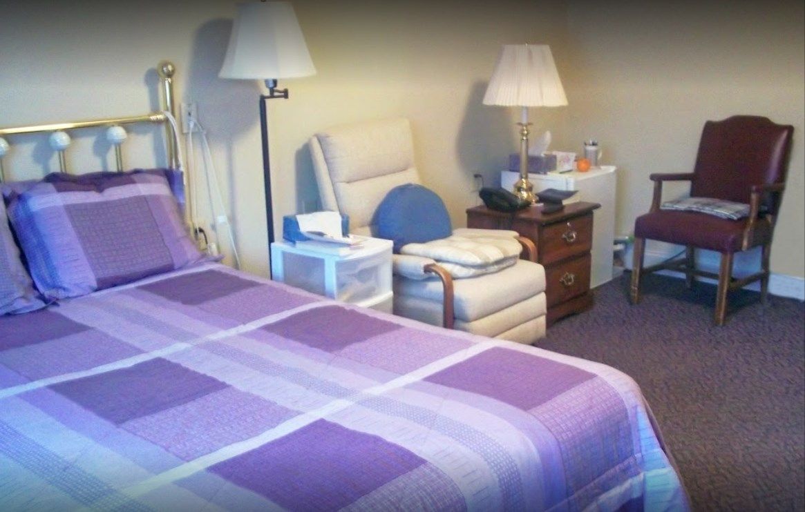 Senior living community bedroom at Cambridge House featuring a lamp, chair, and bed.