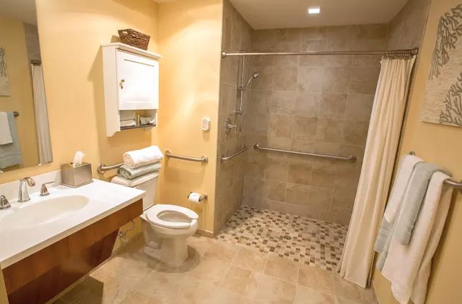 Indoor view of a bathroom with sink, shower, and toilet at Symphony at St. Augustine senior living community.