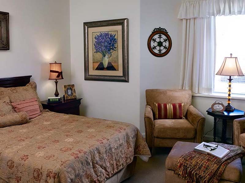 Senior living room at Atria Summit Ridge with bed, chair, table lamp, book, art painting, and decor.