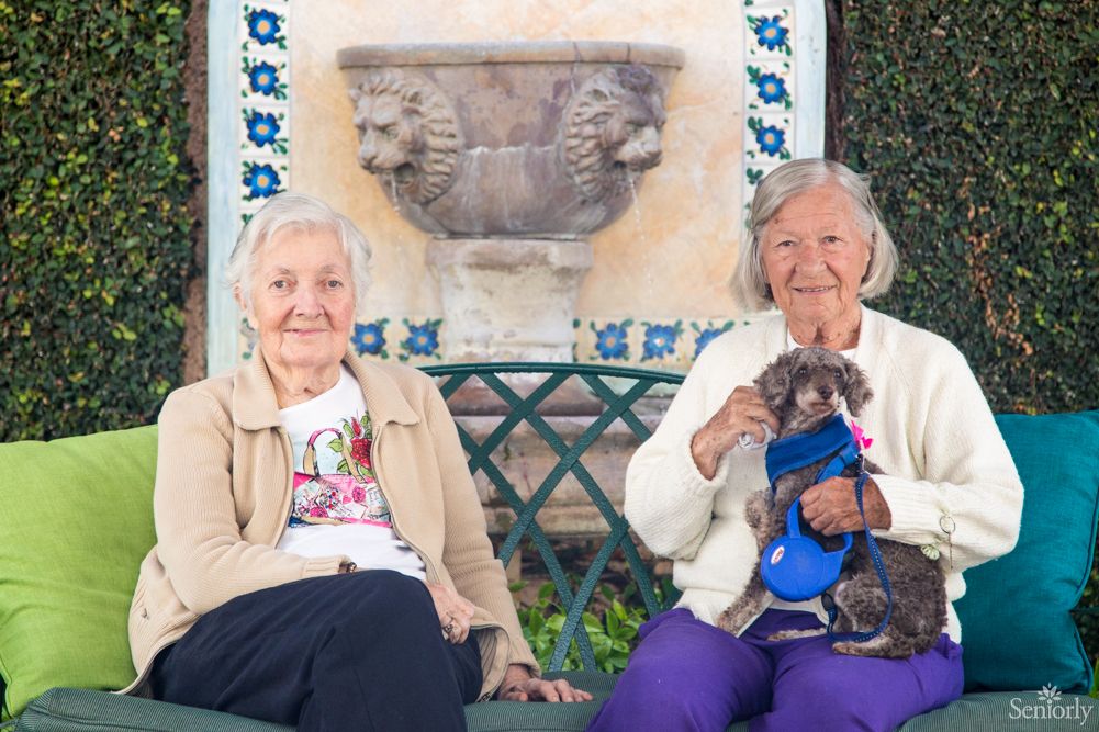 Elderly woman in knitwear sitting on couch with pet dog at Burlingame Villa senior living community.