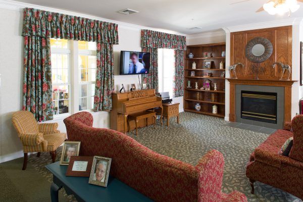 Senior living room interior at Juniper Village, featuring a piano, TV, fireplace, and cozy decor.